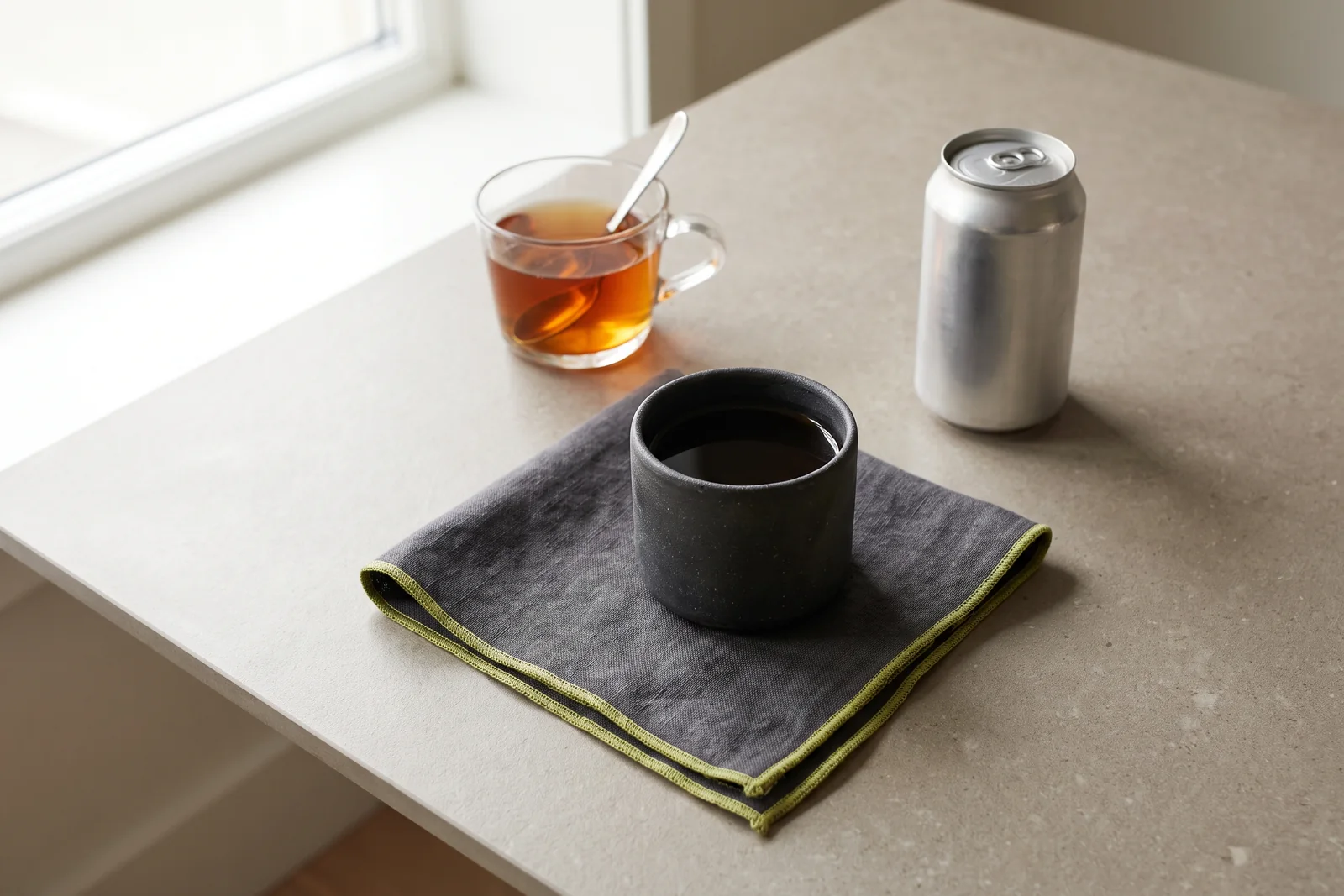 A cup of black coffee, a glass of tea and a plain metal drink can on a grey tabletop.