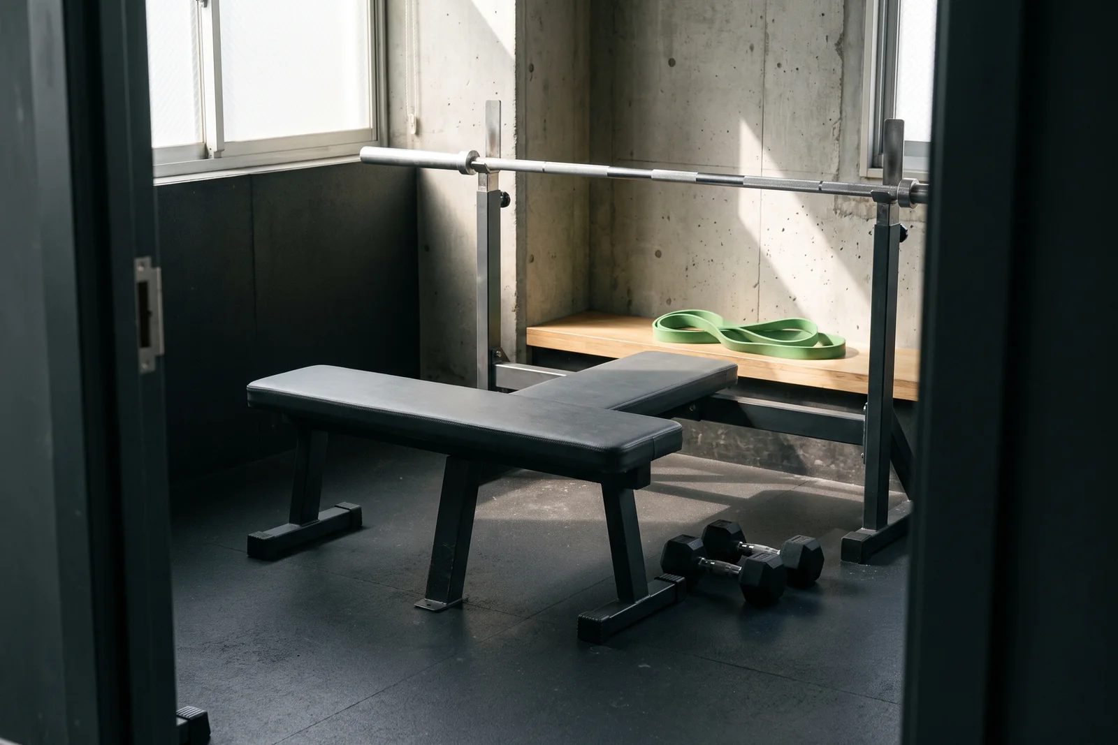 A workout bench, racked barbell and small dumbbells in an empty concrete-walled gym.