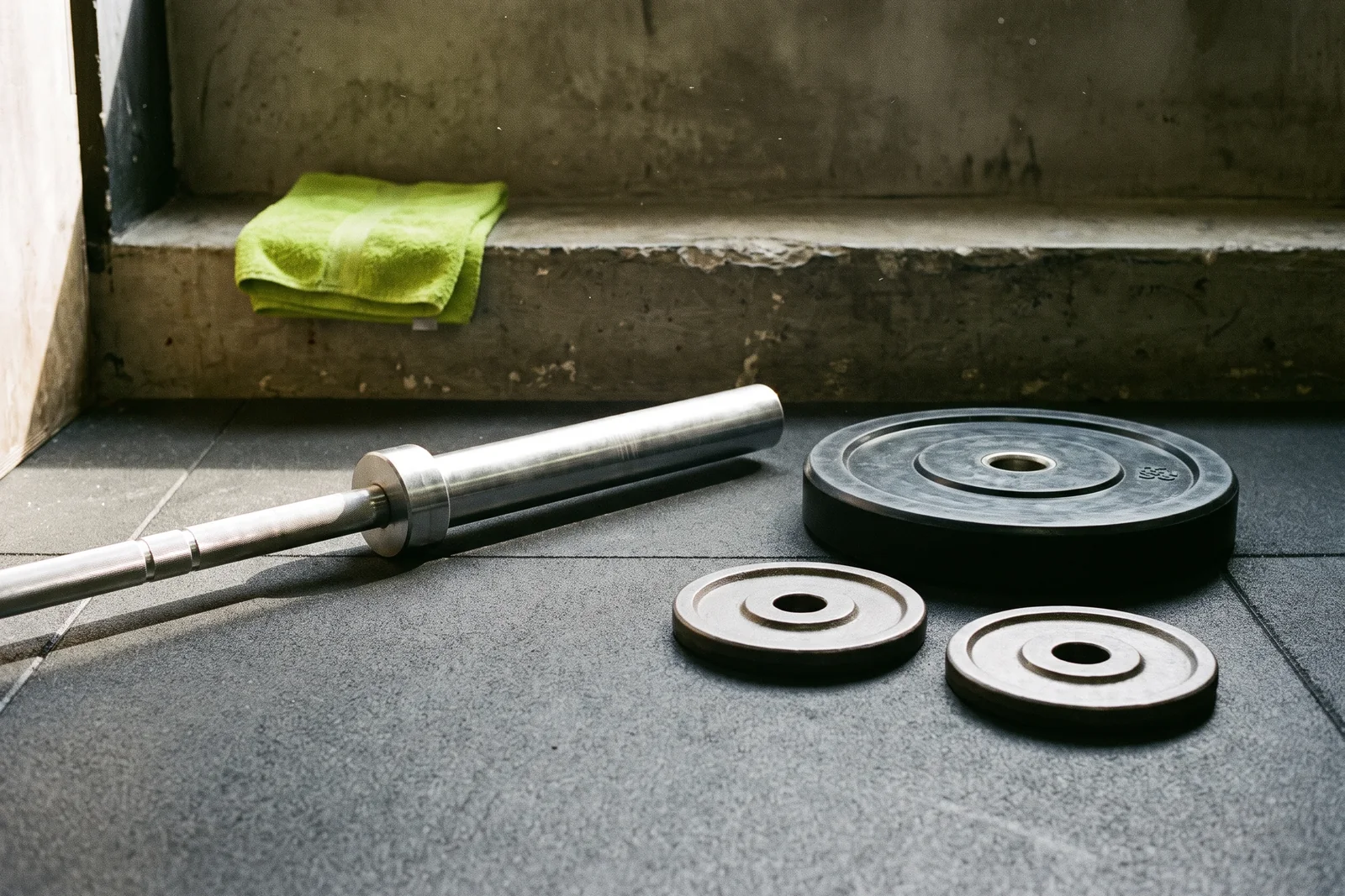 A bare barbell sleeve and three loose weight plates on a rubber floor beneath a green towel.
